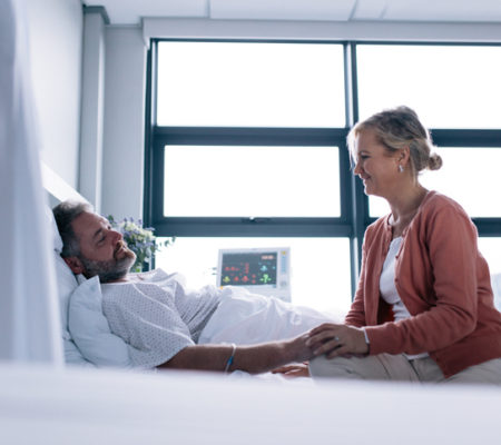 Woman visiting husband in hospital. Female talking with male patient lying in hospital bed.